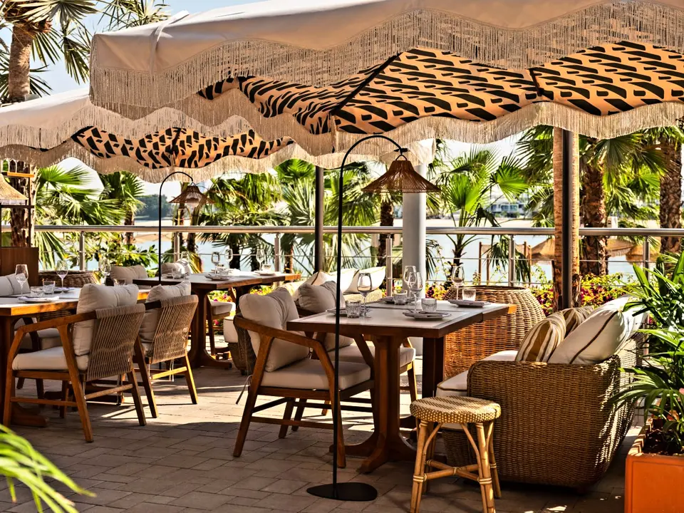 Beachfront restaurant terrace furnished with teak dining chairs, marble topped tables and rattan lounge seating under fringed parasols